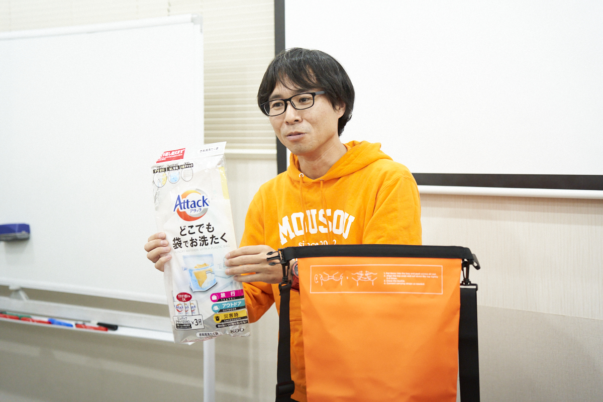 オレンジ色の服を着た男性が、洗濯用の袋を手に持ち、大きなオレンジのバッグの横で説明しています。