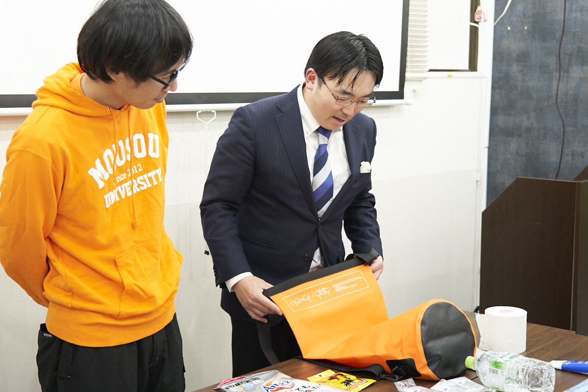 スーツ姿の高荷さんとオレンジのパーカーを着た地主さんが、テーブル上のオレンジ色のバッグとアイテムを確認しています。
