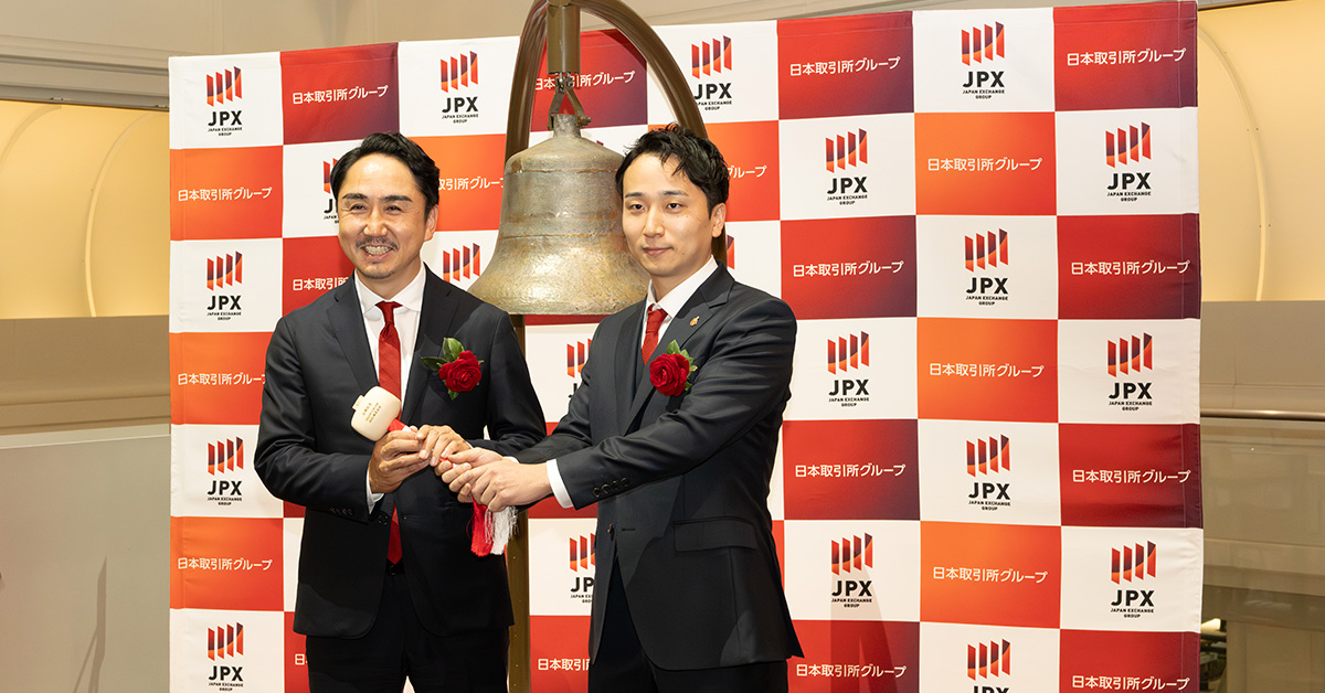 スーツを着た堀江と出澤性が、JPXのロゴが入った背景の前で、鐘を鳴らすためのハンマーを一緒に持っています。