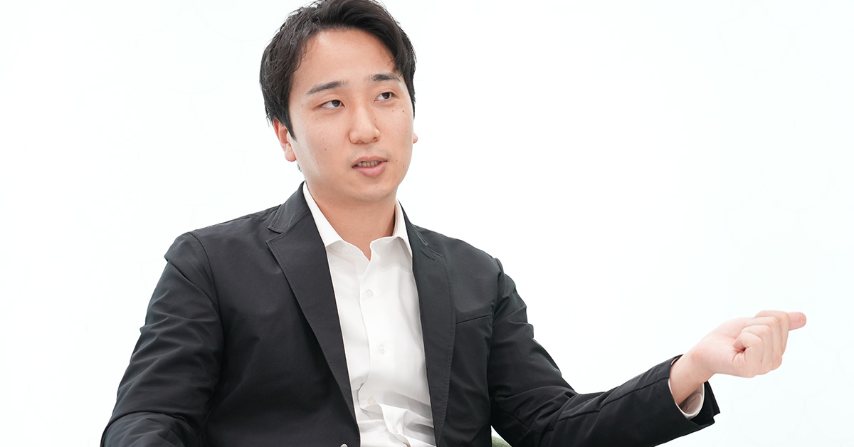 黒いジャケットに白いシャツを着た堀江が、手を広げて話をしている様子です。背景は淡い色です。
