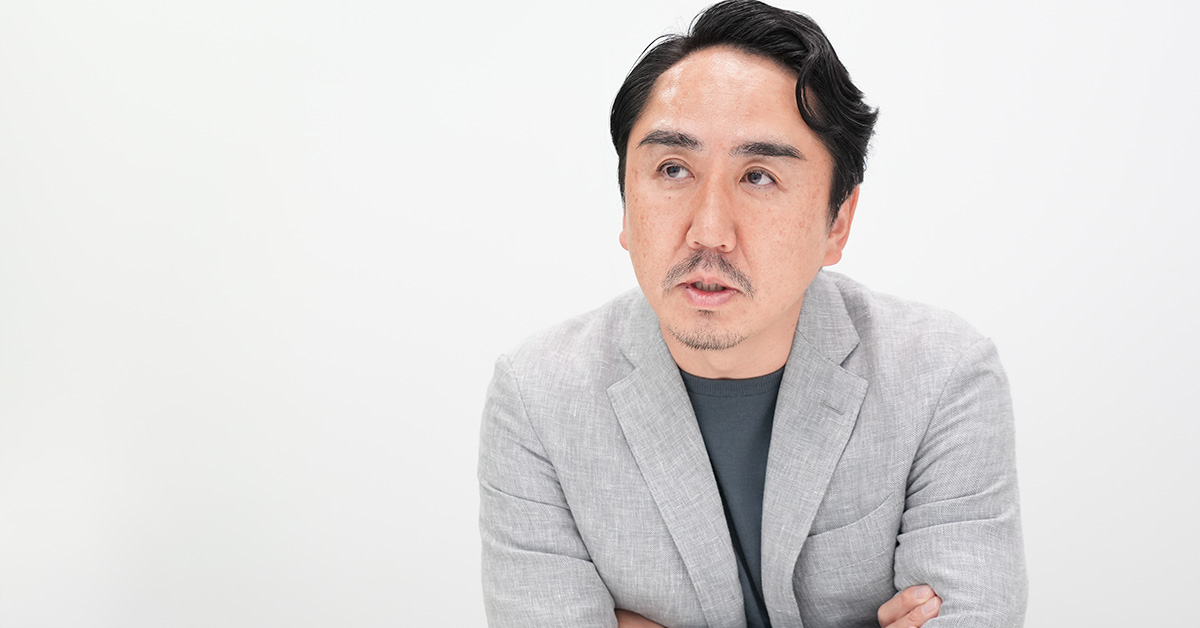 グレーのジャケットを着た出澤が腕を組み、何かを考えながら話している様子です。背景は白です。