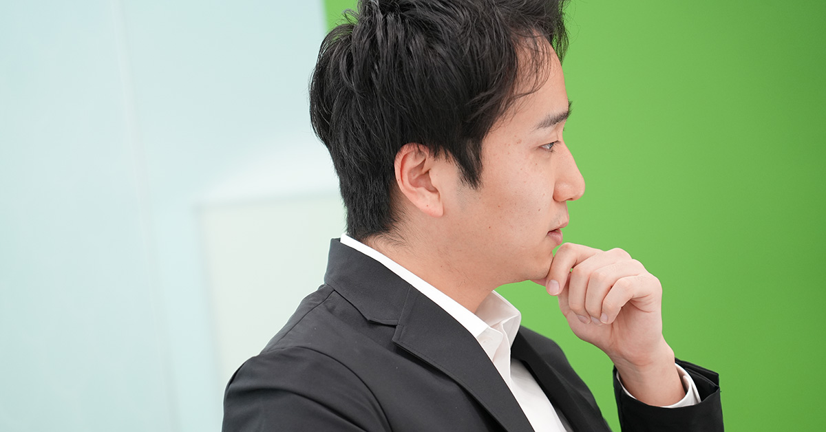 黒いジャケットに白いシャツを着た堀江が、手を顎に当てて考えている様子です。