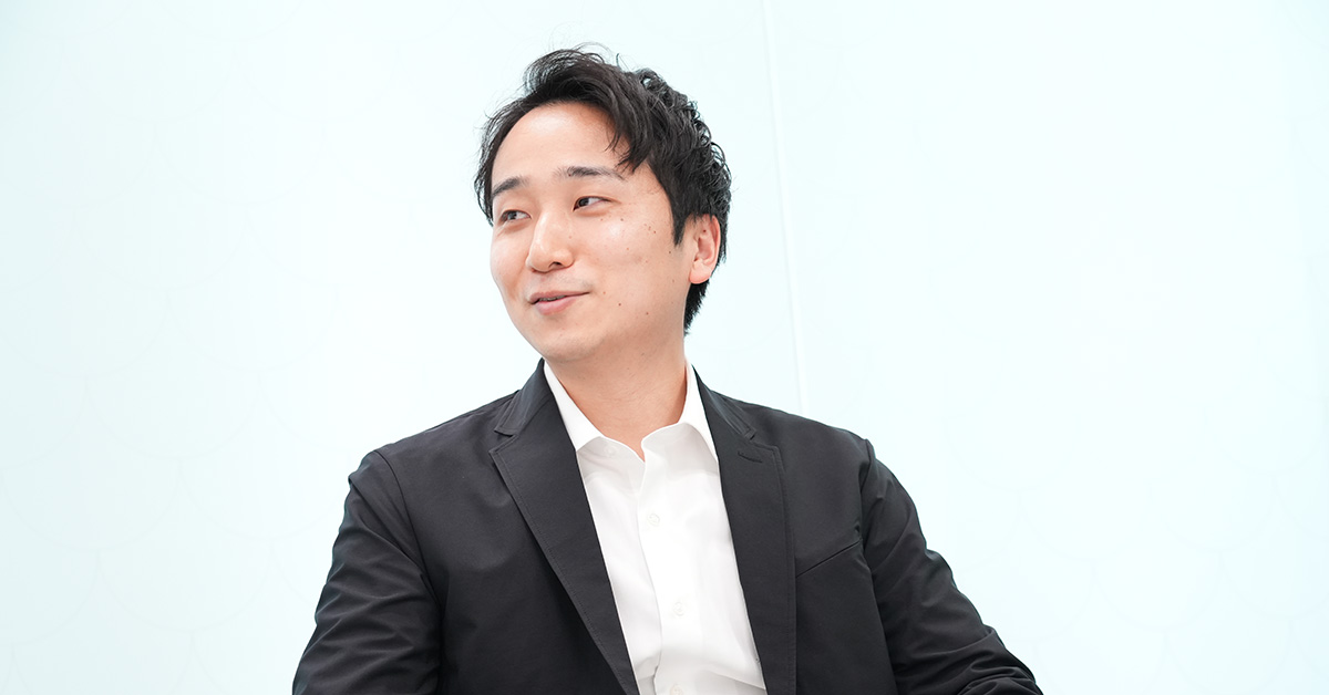黒いジャケットに白いシャツを着た堀江が、淡い青の背景の前で微笑んで横を向いている様子です。