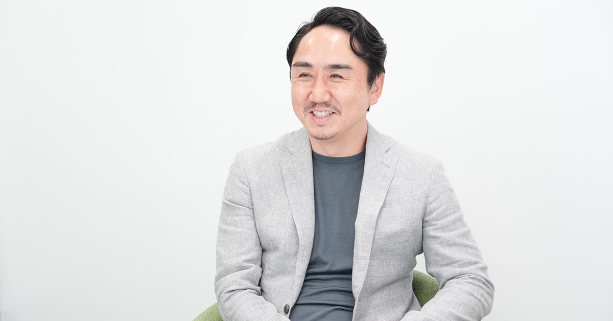 グレーのジャケットを着た出澤が、白い背景の前で椅子に座り、笑顔でリラックスした様子です。