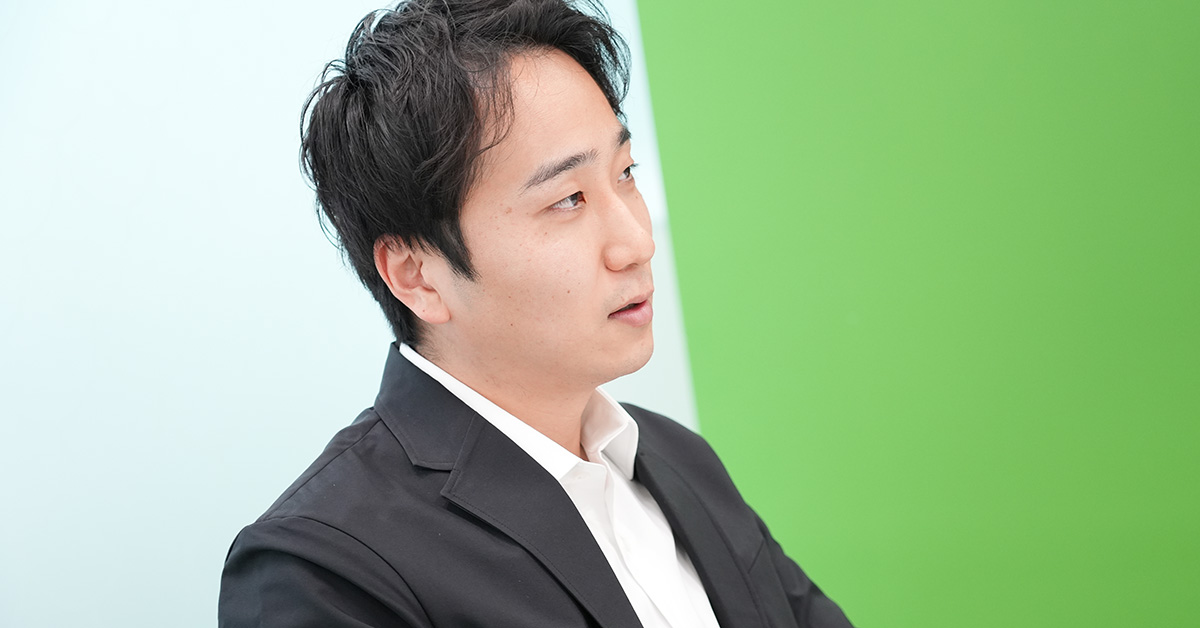 黒いジャケットに白いシャツを着た堀江が、横を向いて話している様子です。背景は明るい緑と青です。