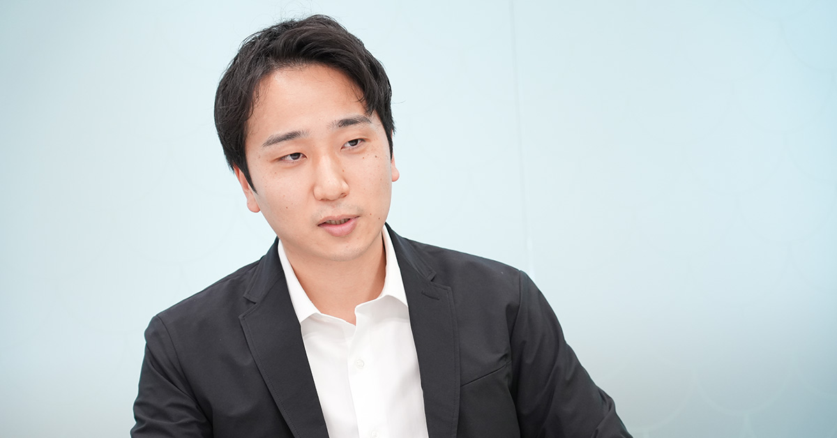 黒いジャケットに白いシャツを着た堀江が、淡い青の背景の前で話をしている様子です。