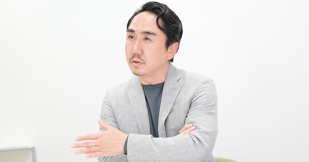 グレーのジャケットを着た出澤が、白い背景の前で腕を組んで話している様子です。