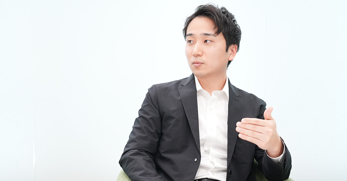 黒いジャケットに白いシャツを着た堀江が、白い背景の前で手を使いながら話している様子です。