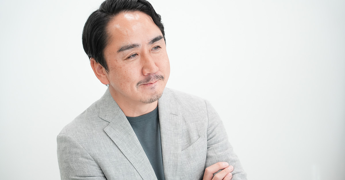 グレーのジャケットを着た出澤が、腕を組んで穏やかな表情で遠くを見ている様子です。