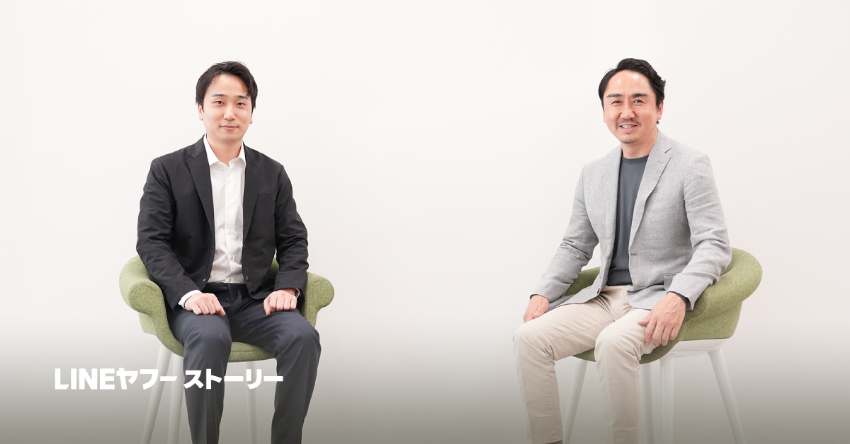 二人の男性、堀江と出澤が、白い背景の前で緑の椅子に座って微笑んでいます。左の堀江は黒いジャケット、右の出澤はグレーのジャケットを着ています。