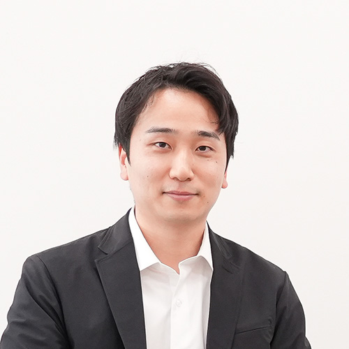 黒いジャケットに白シャツを着た堀江が、白い背景でリラックスした感じでカメラを見ています。