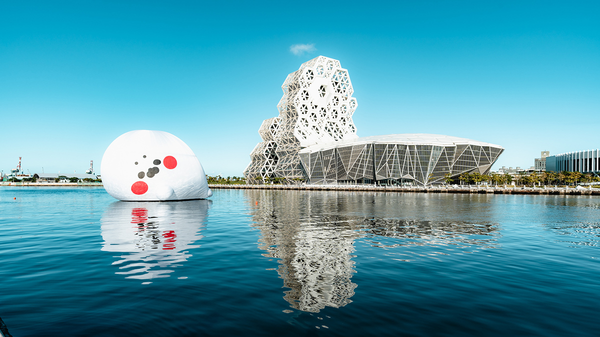 水辺に近未来的なデザインの建物と、大きな白いキャラクターのバルーンが浮かんでいる風景です。青空が広がり、穏やかな印象を与えます。