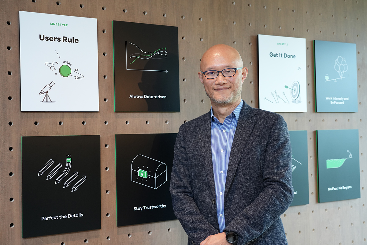 壁に掲示されたLINEの理念パネルの前で、スーツ姿の男性が微笑んで立っている様子です。