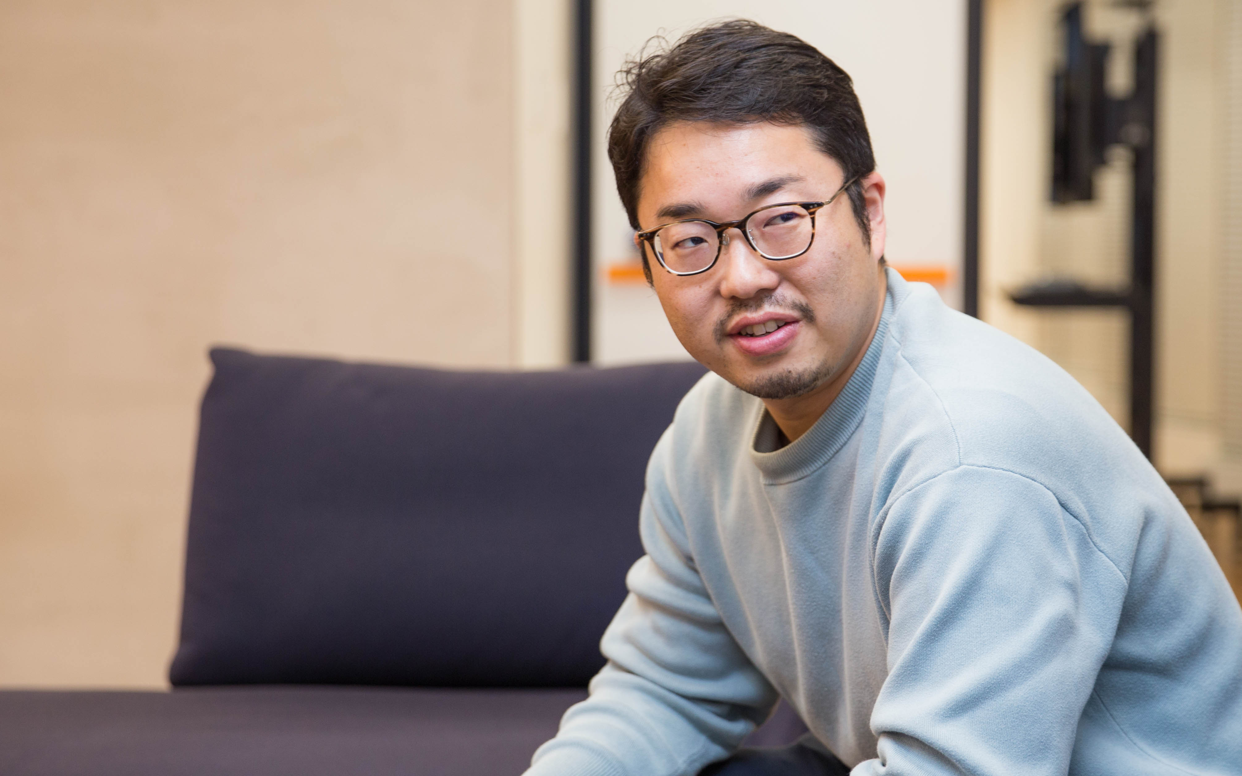眼鏡をかけた男性、三宅がソファに座り、リラックスした表情で横を向いています。