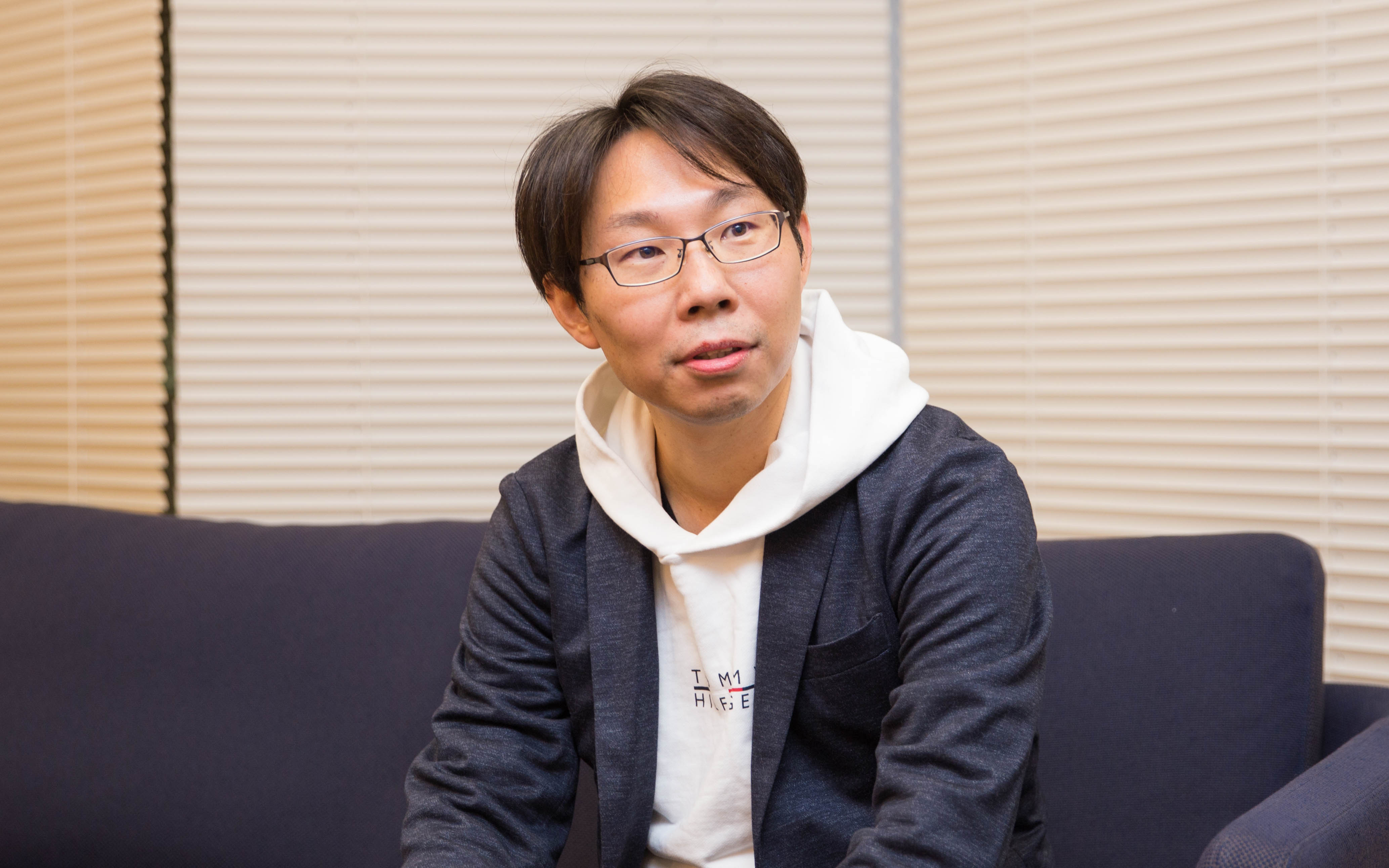 眼鏡をかけた男性、長妻がソファに座り、リラックスした様子で話しています。背景にはブラインドが見えます。