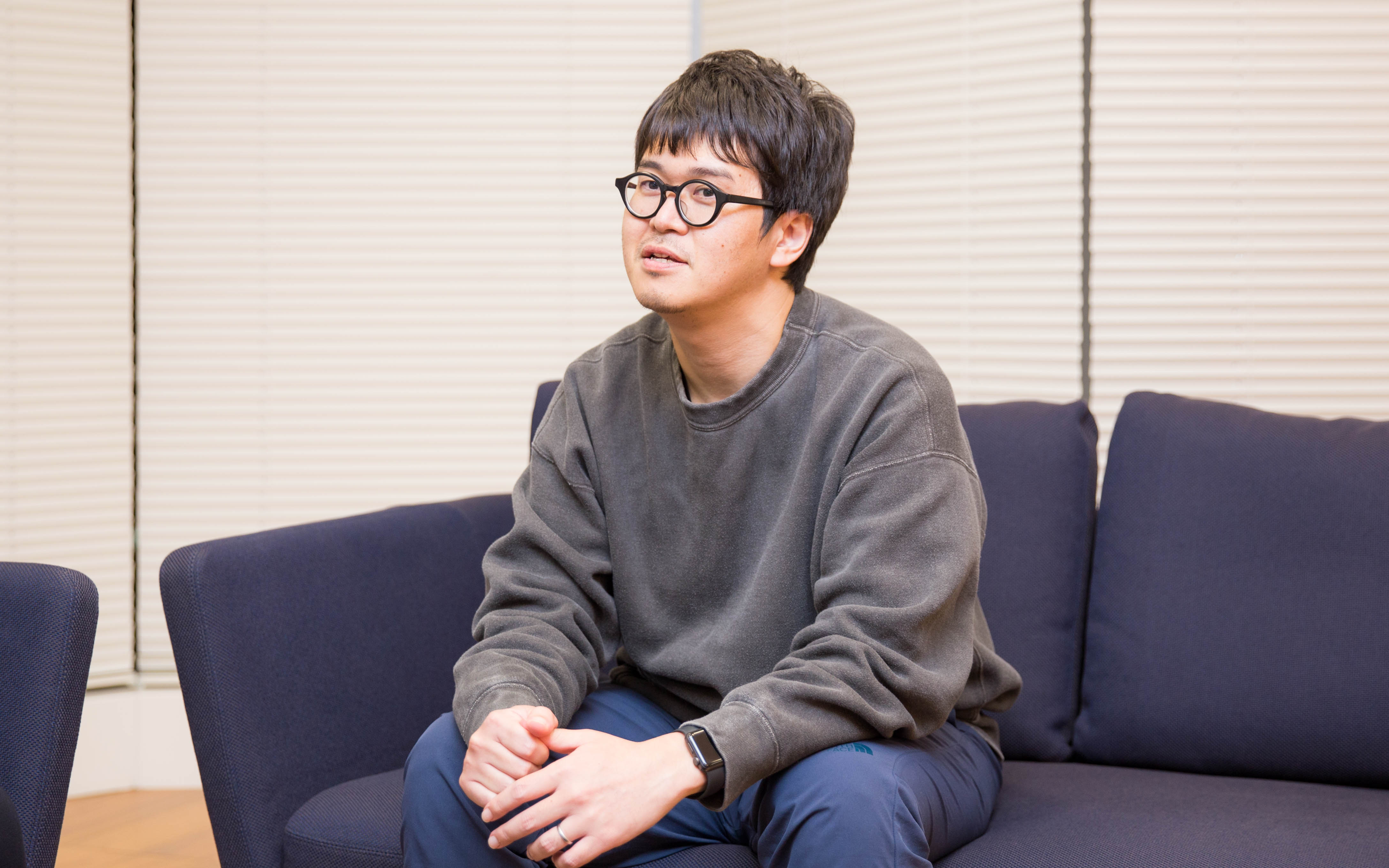 眼鏡をかけた男性、西丸がソファに座り、少し前傾して話を聞いています。背景にはブラインドが見えます。