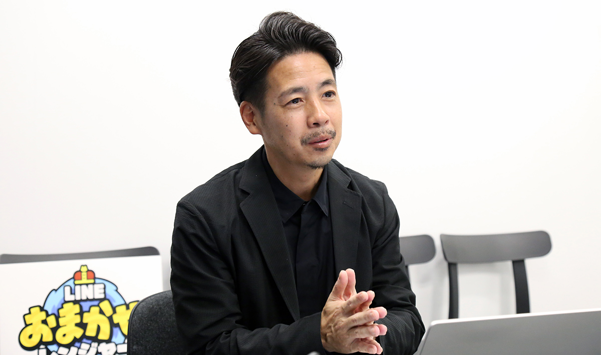 黒い服を着た男性が、室内で手を組みながら話をしている様子。「LINE おまかせレンジャー」のロゴが背景にある。