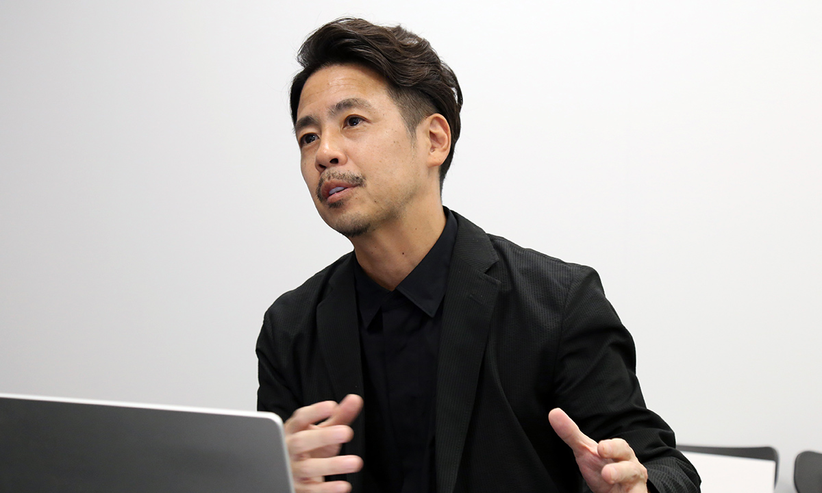 黒い服を着た男性が室内でジェスチャーを交えて話をしている。背景には白い壁と椅子が見える。