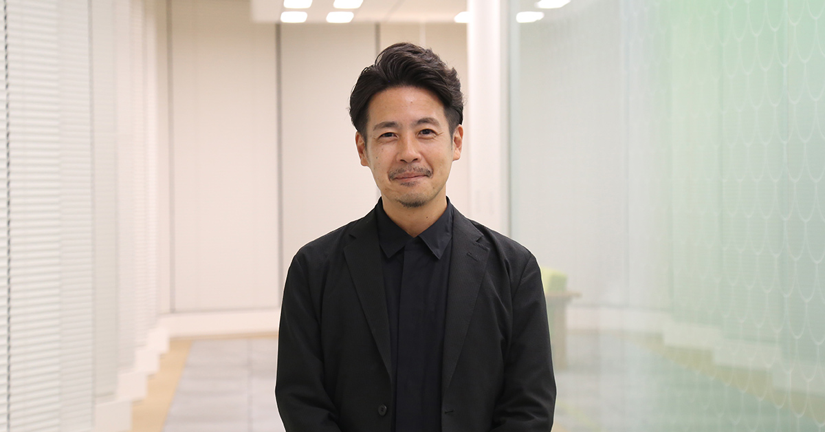 黒い服を着た男性が、明るい室内の廊下で穏やかな表情で立っている。背景に白いパーティションが見える。