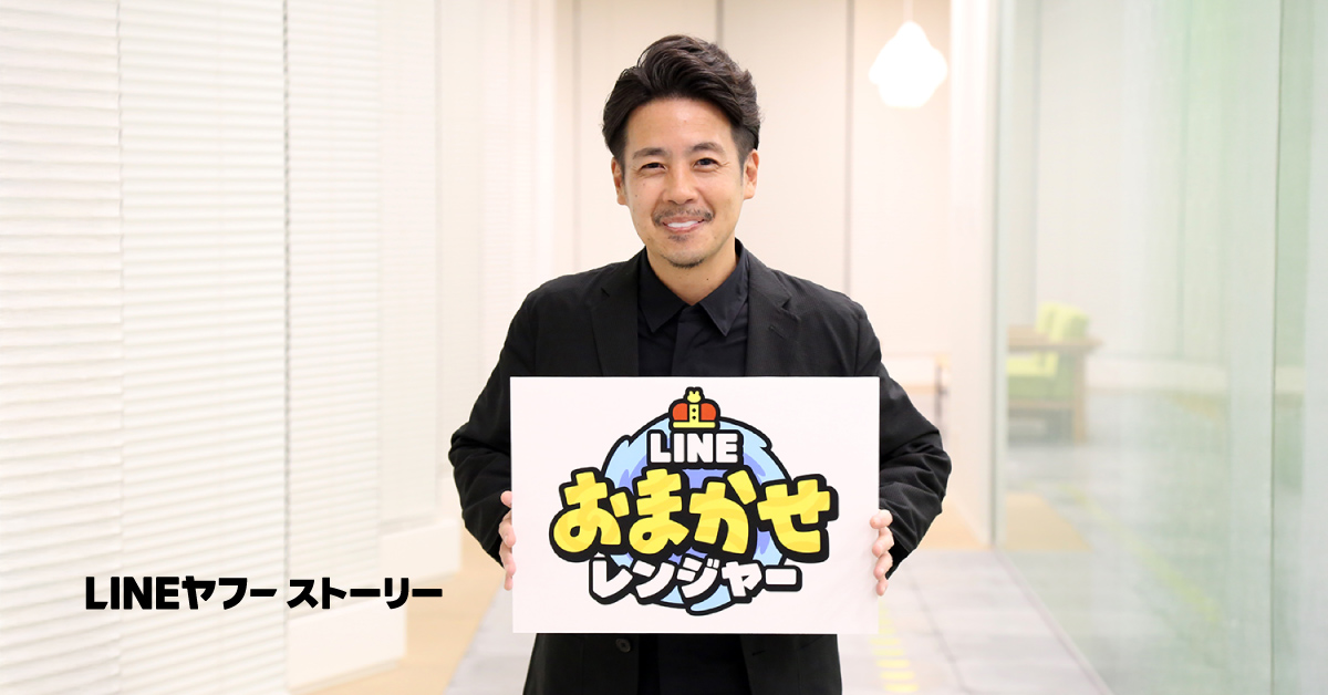 室内で黒い服を着た男性が「LINE おまかせレンジャー」と書かれた看板を持って笑顔を浮かべている