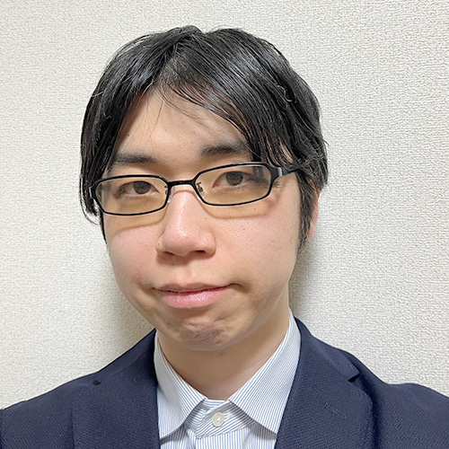 スーツを着た大神が、メガネをかけて正面を見つめています。背景は白です。