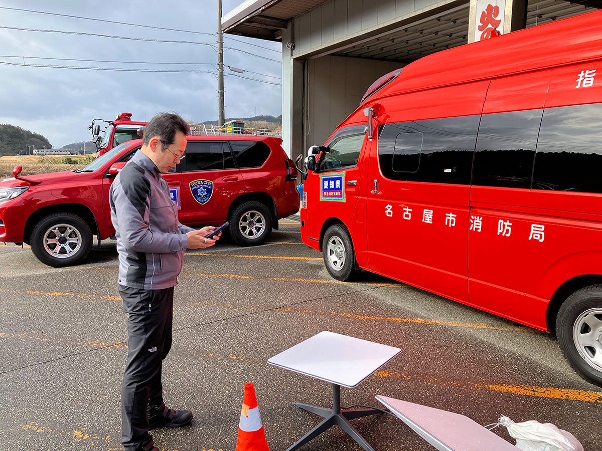 赤い消防車と車が停まっている駐車場で、男性がスマートフォンを操作しています。名古屋市消防局の文字が見えます