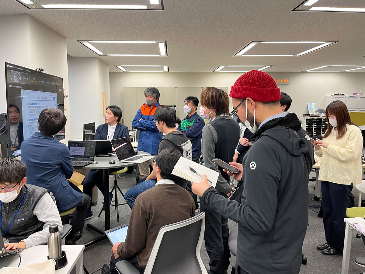 オフィスでのミーティング風景。大勢の人がスクリーンを見ながらディスカッションしています