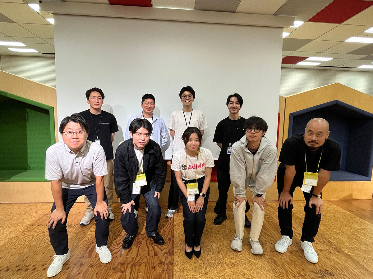 9人の男女が室内でポーズをとっている集合写真です。背景にはカラフルな壁や床があり、カジュアルな雰囲気が漂っています。