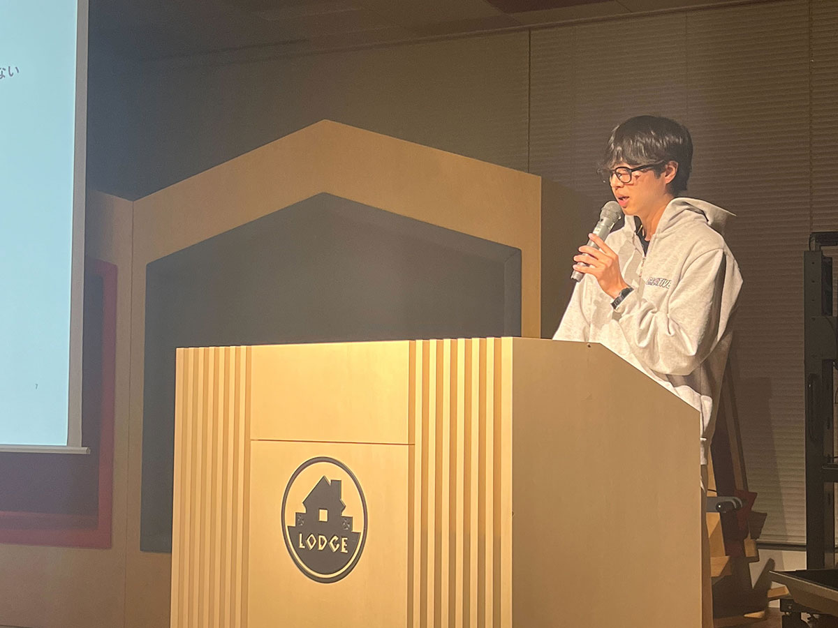 フード付きのパーカーを着た男性が、マイクを持って講演している写真です。講演台には「LODGE」と書かれており、プレゼンテーションの一場面です。