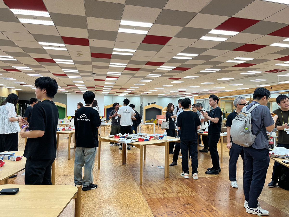 オフィスのような場所で、多くの人々が立ちながら交流している写真です。テーブルには食べ物や飲み物が置かれ、カジュアルな雰囲気のイベントが行われています。
