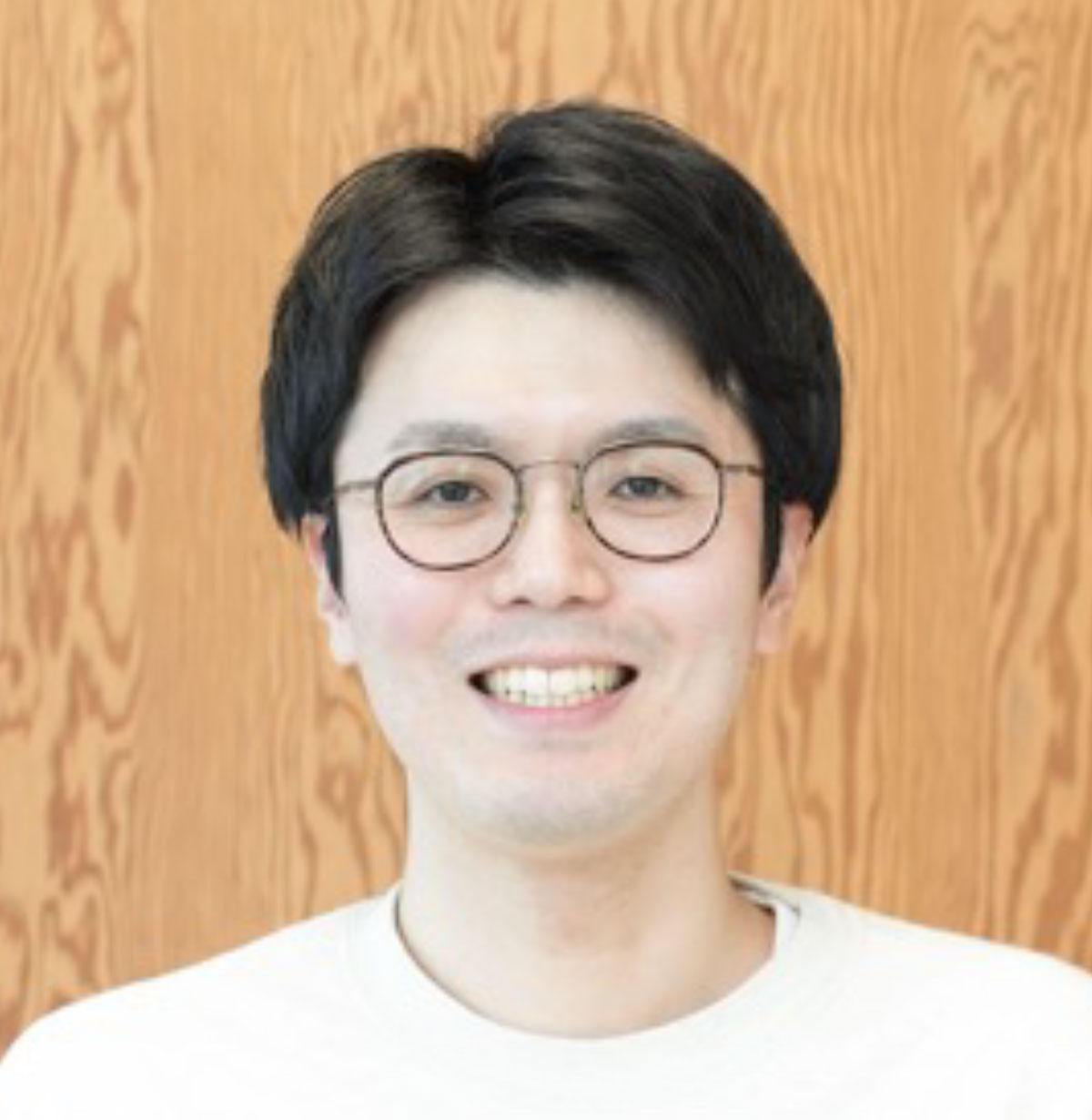 眼鏡をかけた亀岡が木目の壁を背景に笑顔を見せている写真です。カジュアルな雰囲気が感じられます。