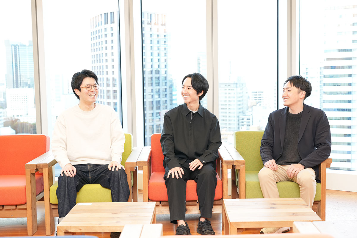3人の男性が窓際の椅子に座って談笑している写真です。背景には都市のビル群が広がり、和やかな雰囲気が感じられます。