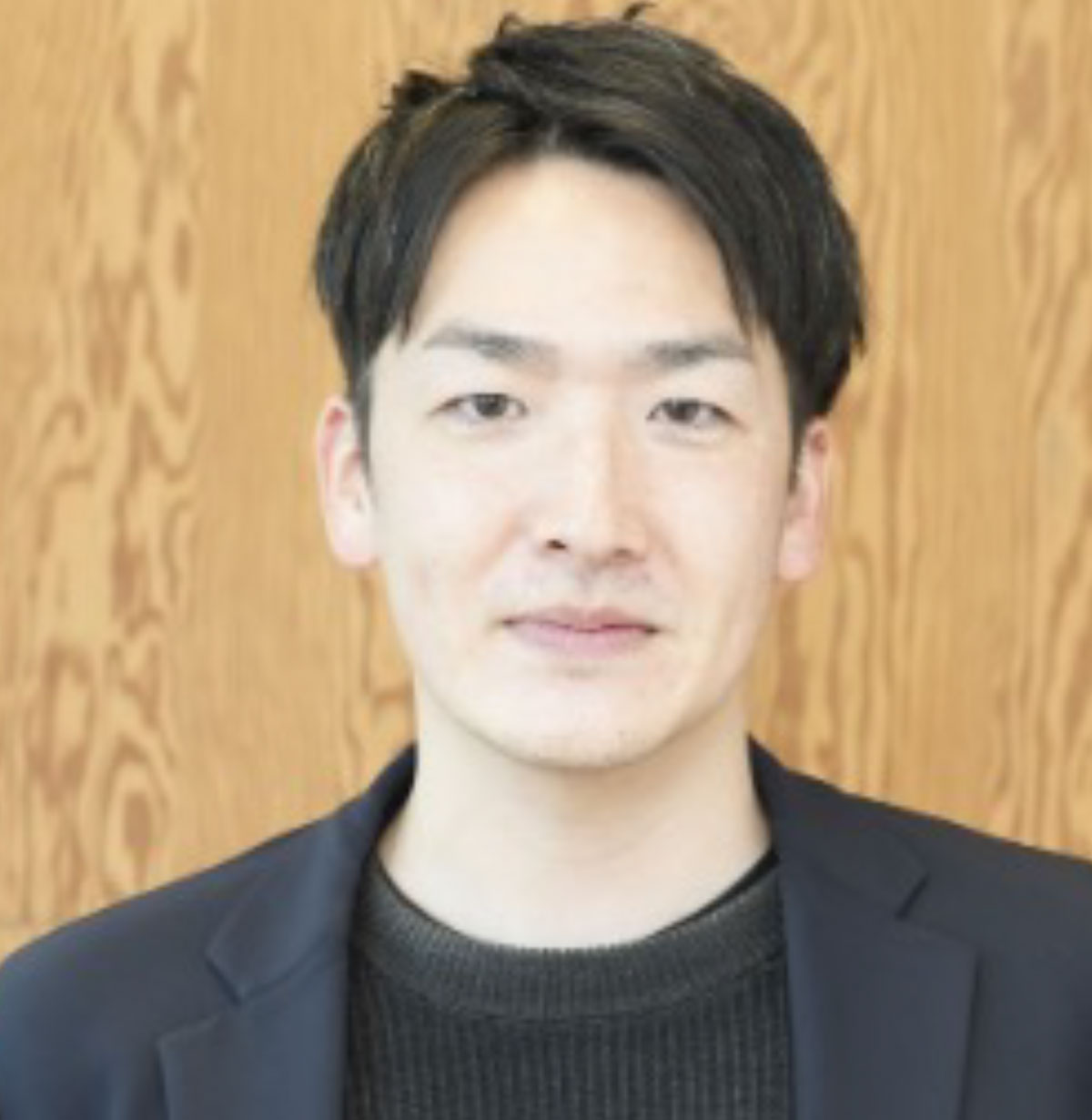 黒いジャケットを着た内丸が木目の壁を背景に立っている写真です。落ち着いた表情でカメラを見ています。