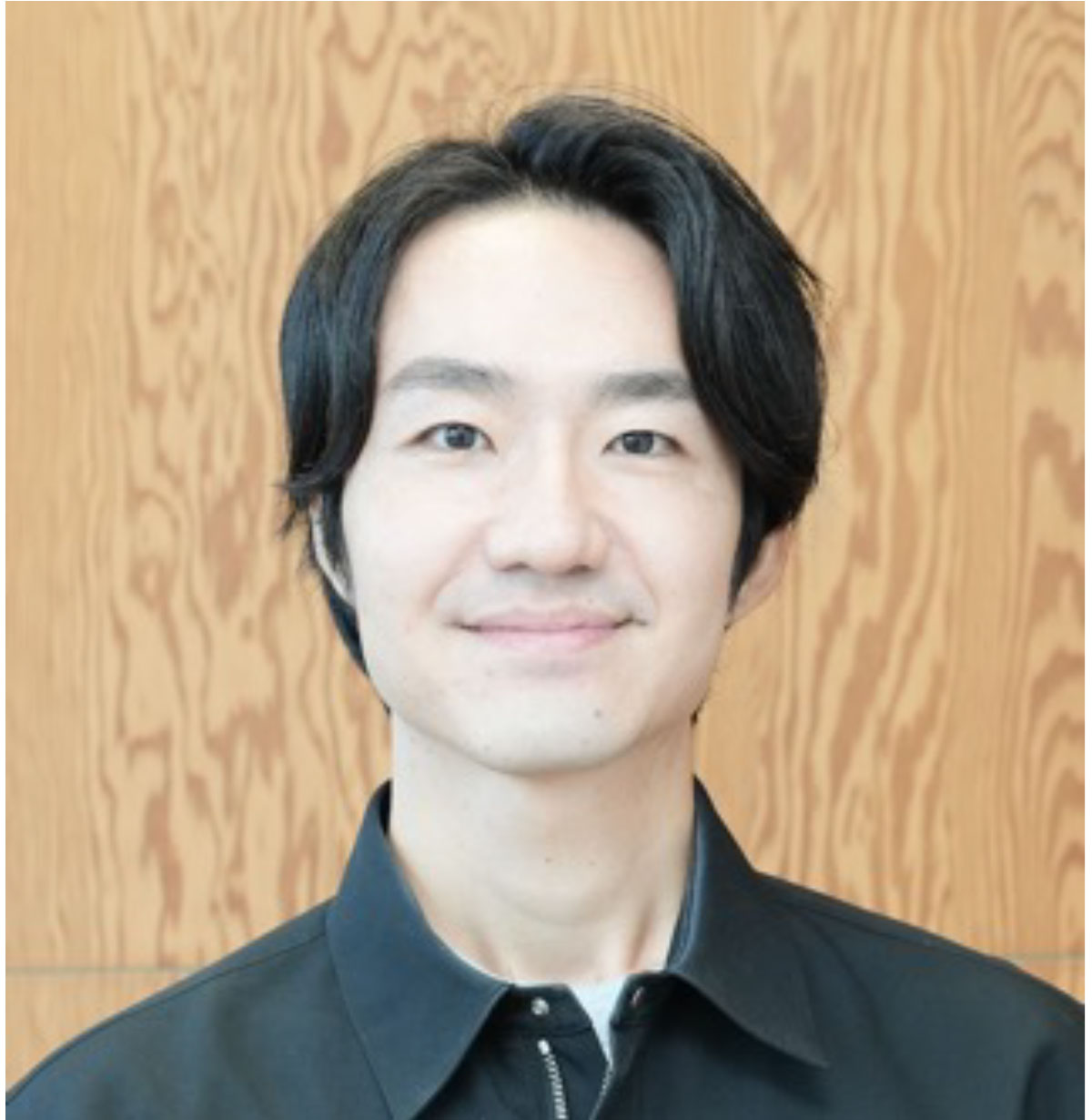 黒いシャツを着た湯田が木目の壁を背景に微笑んでいる写真です。シンプルで落ち着いた雰囲気があります。