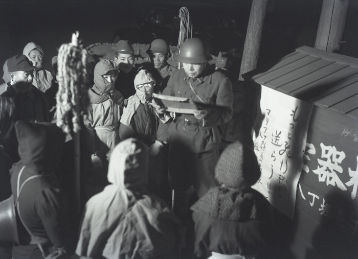 「もっと飛行機を送ろう」の貼り紙の下で実施される防空訓練＝東京都中央区八丁堀で 1944 年 3 月